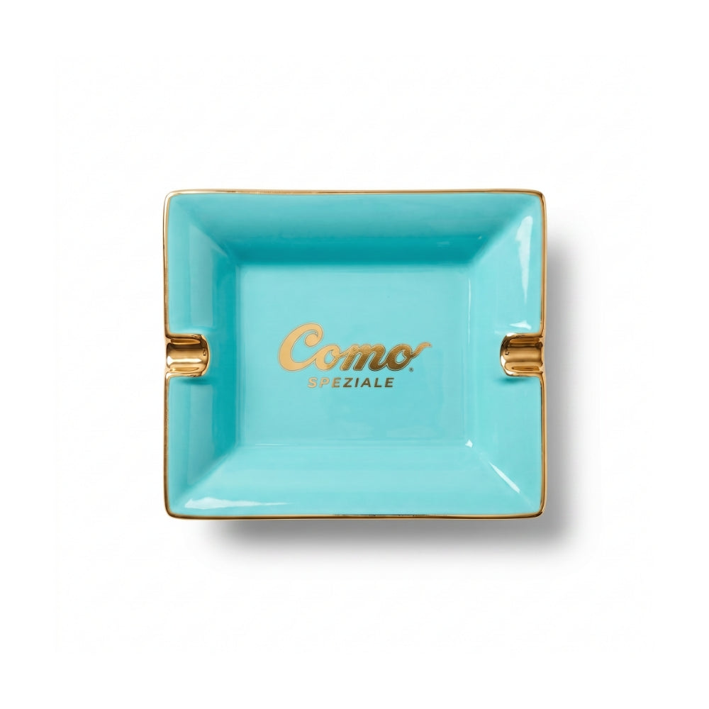 A turquoise blue square ceramic ashtray with gold trim and gold-accented side slots, featuring the 'Como Speziale' logo printed in gold in the center, shown on a white background.
