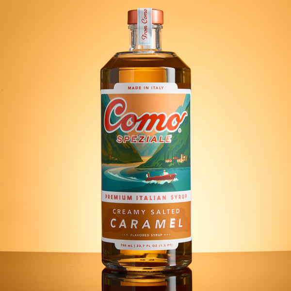A bottle of Como Speziale Creamy Salted Caramel syrup, standing on a reflective surface against a warm golden-yellow gradient background.