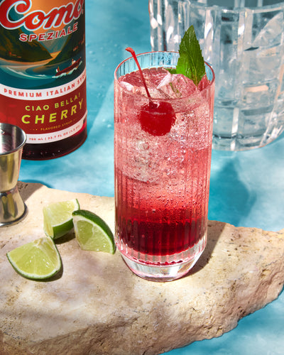 A tall glass of a pink-red cocktail with ice, garnished with a cherry and mint leaf, set on a stone slab with lime wedges. A bottle of Ciao Bella Cherry and a jigger are visible in the background against a bright blue backdrop.