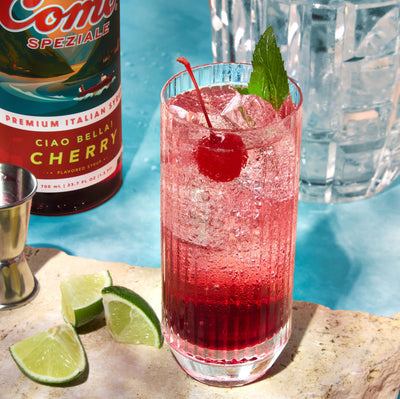 A tall glass of a pink-red cocktail with ice, garnished with a cherry and mint leaf, set on a stone slab with lime wedges. A bottle of Ciao Bella Cherry and a jigger are visible in the background against a bright blue backdrop.