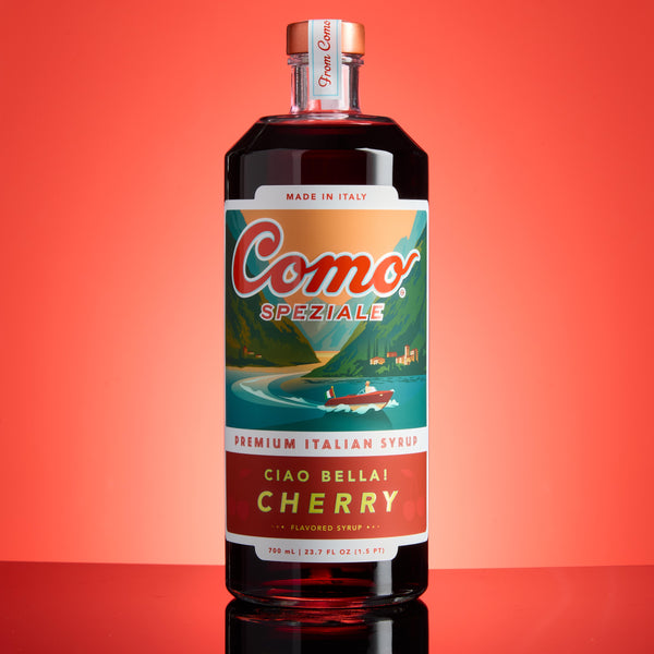 A bottle of Como Speziale Cherry flavored syrup on a reflective red surface against a solid red background. The bottle has a vintage-style label with a boat illustration and the text 'Como Speziale' and 'Ciao Bella Cherry'.