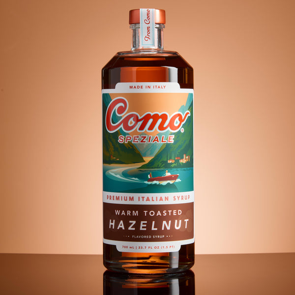 A bottle of Como Speziale Warm Toasted Hazelnut flavored syrup, featuring a brown label with a scenic Italian landscape illustration, set against a warm peach background.