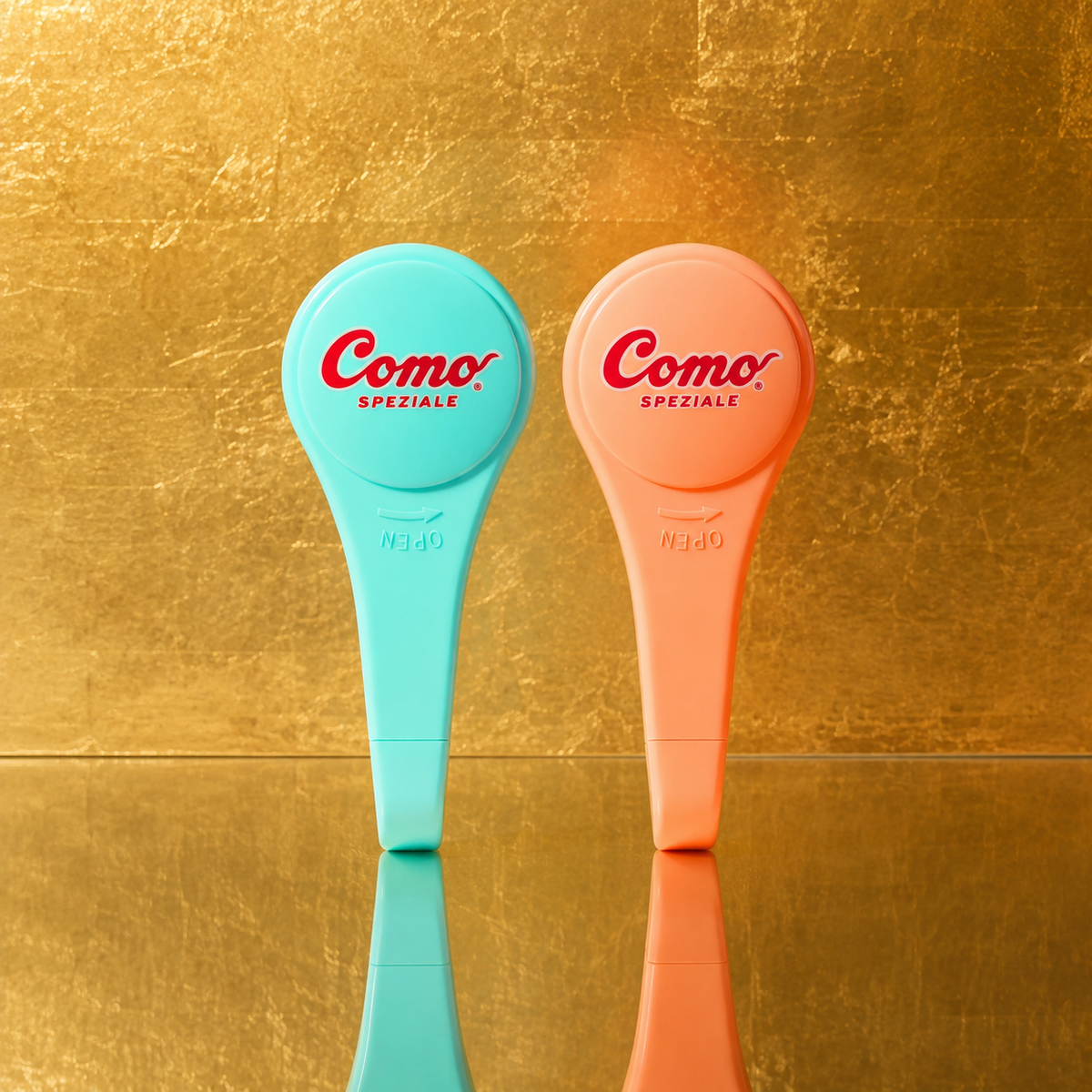 Two pastel-colored plastic syrup pumps stand upright on a reflective gold surface against a textured gold background. The left tool is light aqua blue and the right tool is light peach-pink, both with red 'Como Speziale' branding on the heads.