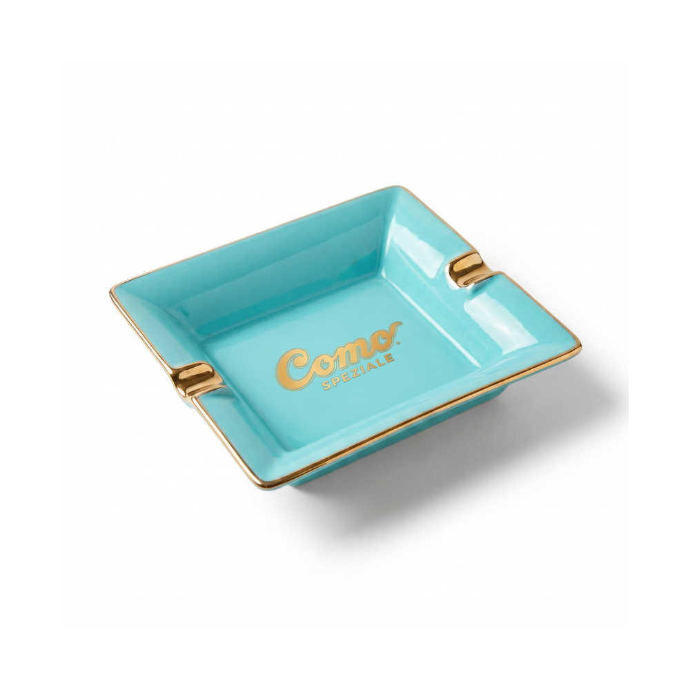 A turquoise blue square ceramic ashtray with gold trim and the 'Como' logo printed in gold on the bottom, set against a plain white background.