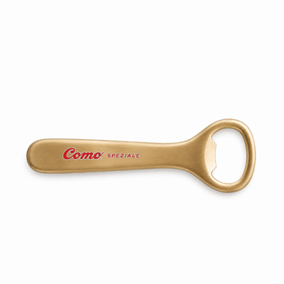 A gold-colored metal bottle opener engraved with 'Como SPEZIALE' in red and black, shown on a plain white background.