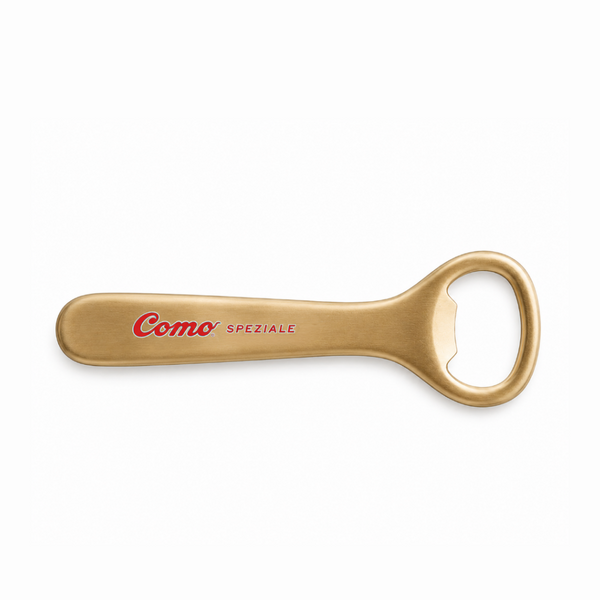 A gold-colored metal bottle opener engraved with 'Como SPEZIALE' in red and black, shown on a plain white background.