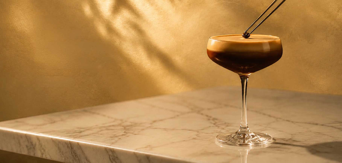A stemmed coupe glass filled with a dark coffee-based drink topped with a light layer of foam, set on a marble surface against a warm golden wall with leaf shadows.