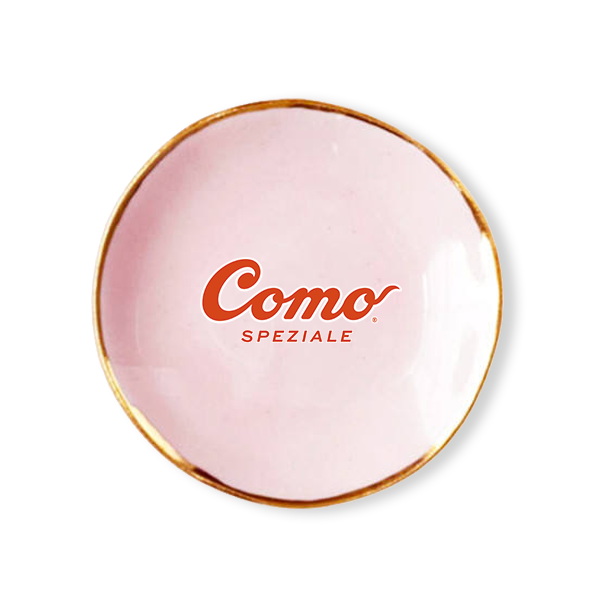 A round pink ceramic plate with a gold rim, featuring the red 'Como Speziale' logo printed in the center, set against a plain white background.